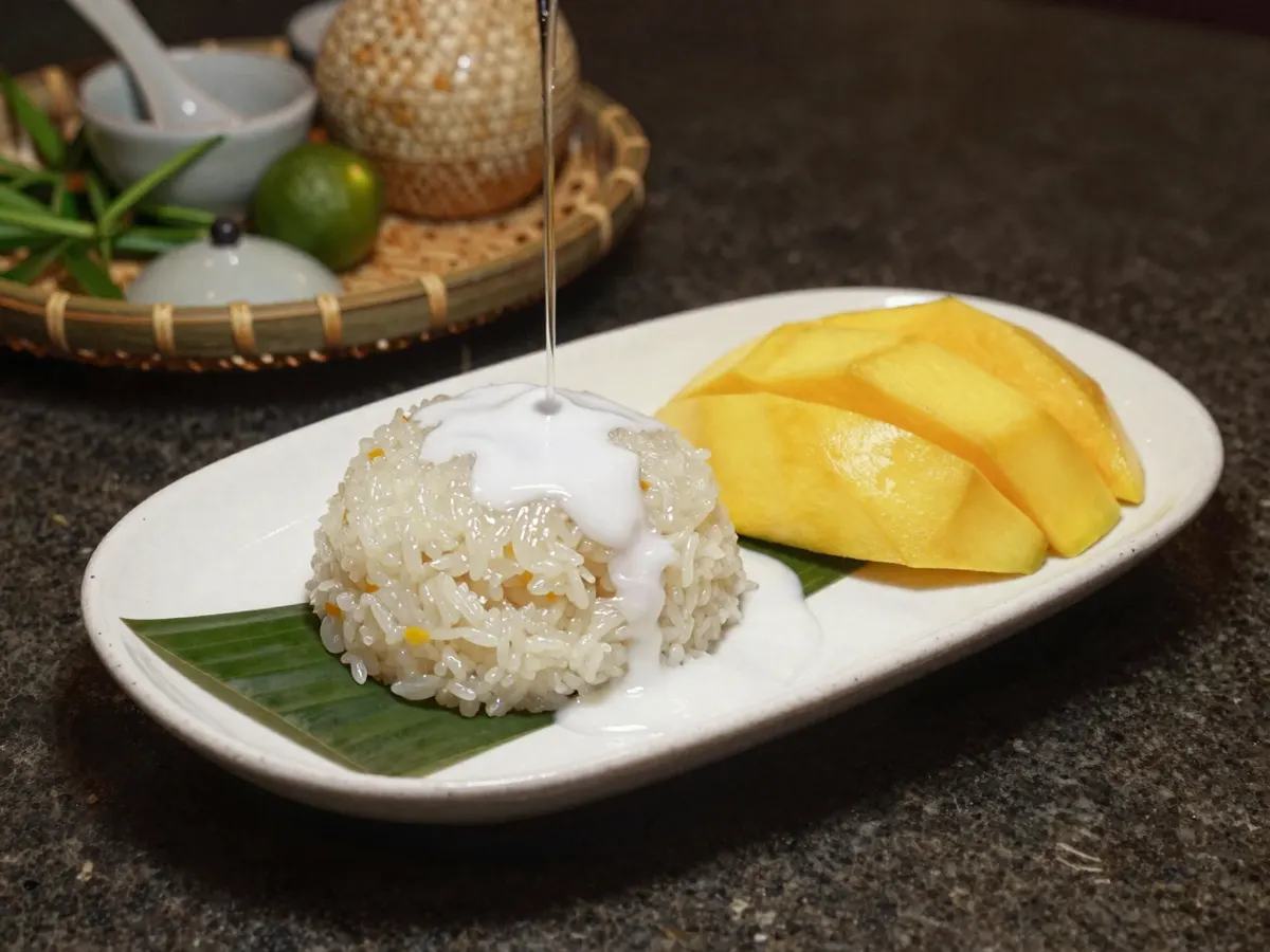 AI-generated photograph: A neat oval mound of glossy white coconut sticky rice with a ribbon of thick coconut cream poured over its top, beside a fan of bright golden yellow mango slices, on a long narrow white porcelain plate, seen from a low three quarter angle, nothing else on the plate