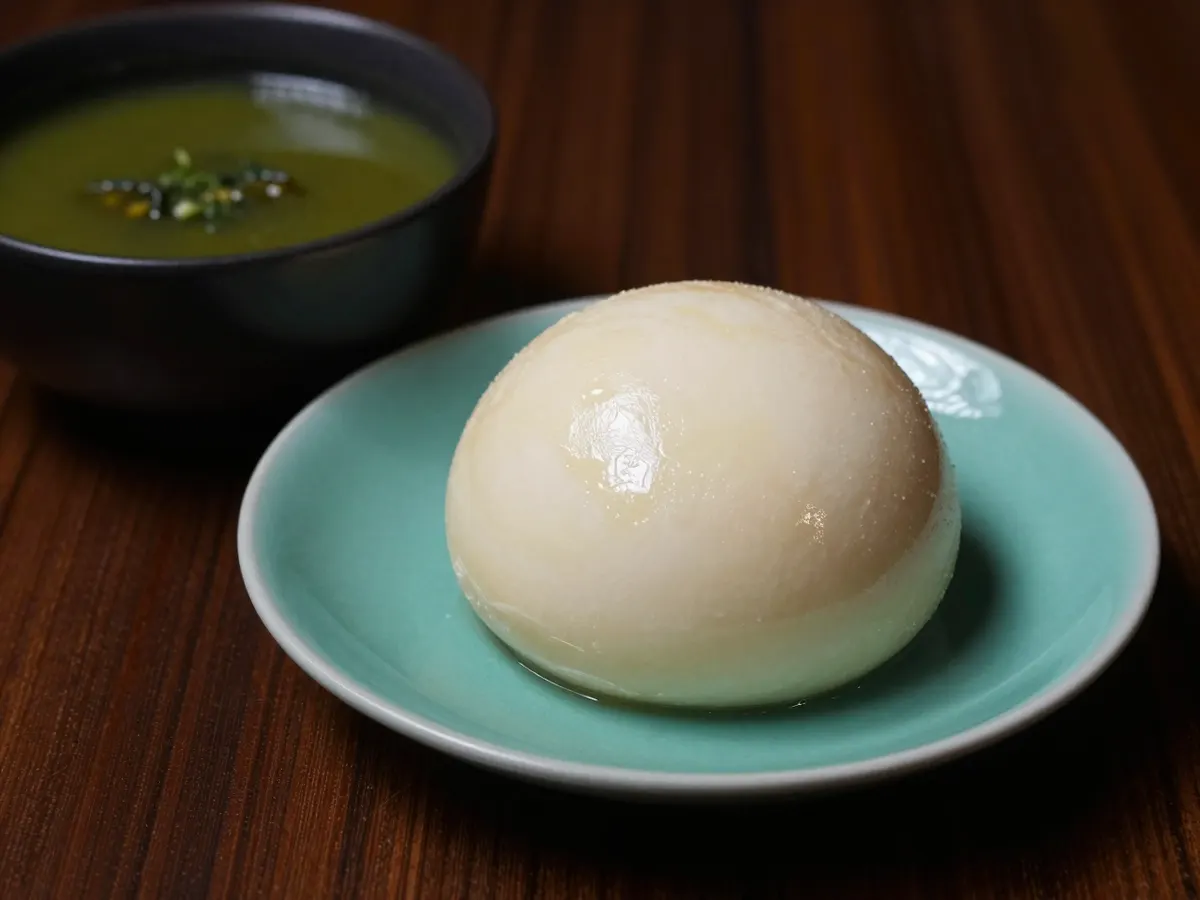 AI-generated photograph: A smooth dome of ivory pounded yam on a small deep celadon plate, its surface glossy and unbroken, beside a dark bowl of green soup just in frame