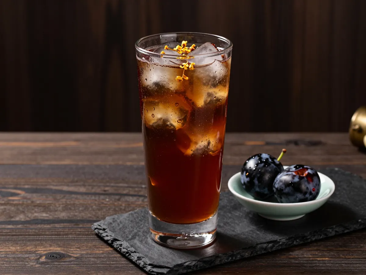 AI-generated photograph: A tall clear glass of deep ruby red plum drink with ice cubes and a few tiny golden osmanthus flowers floating on top, a small dark smoked plum on a saucer beside it, condensation on the glass, seen from the side
